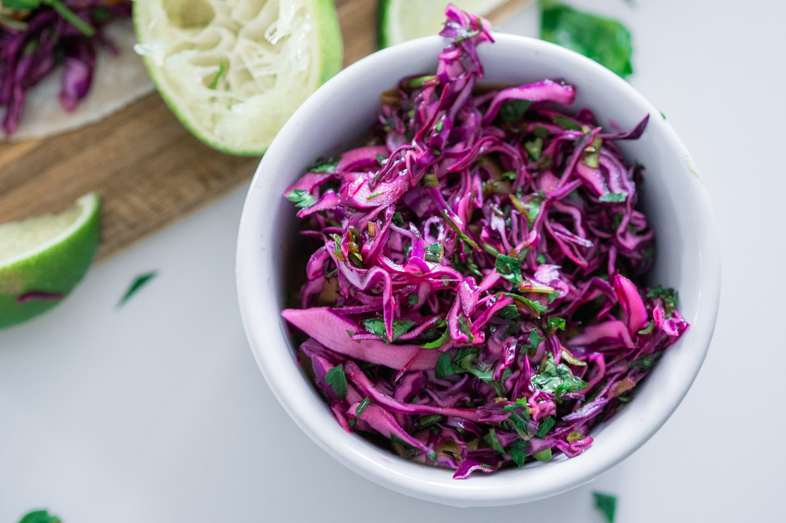 Fresh purple cabbage slaw in a white bowl beside limes. Used to spiced fish tacos.