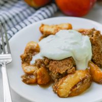 Paleo peach cobbler with coconut whipped cream on a plate with peaches in the background.