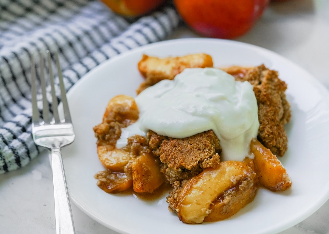Paleo peach cobbler with coconut whipped cream on a plate with peaches in the background.