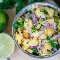 Fresh pineapple salsa in a bowl with limes and parsley in the background.