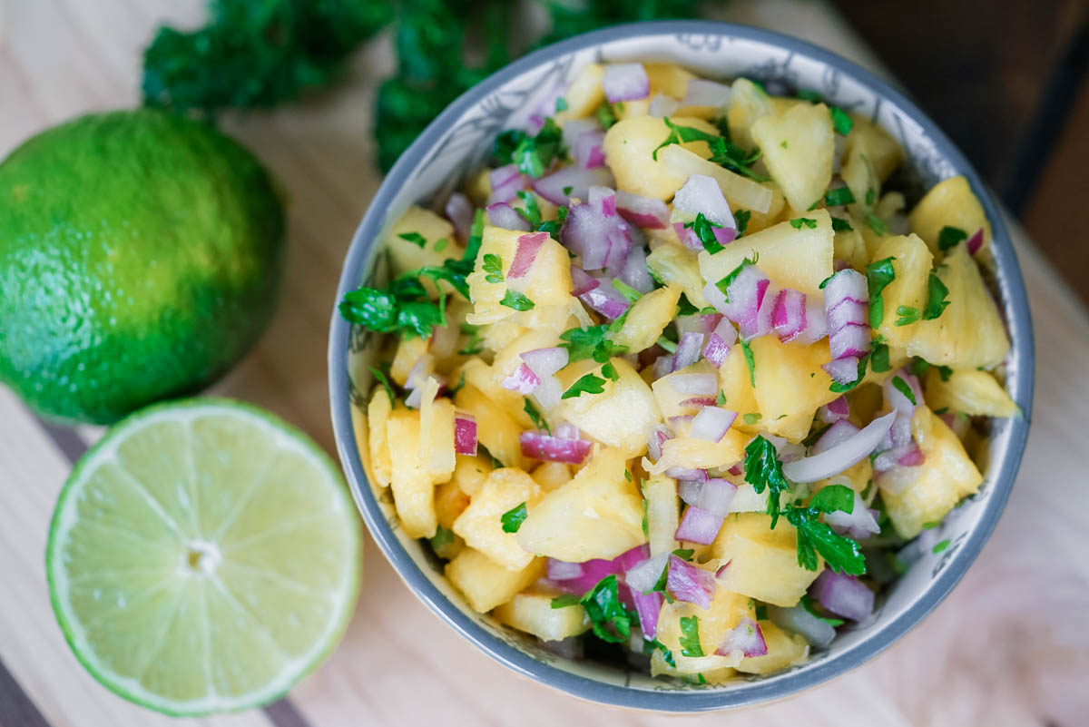 Fresh pineapple salsa in a bowl with limes and parsley in the background.