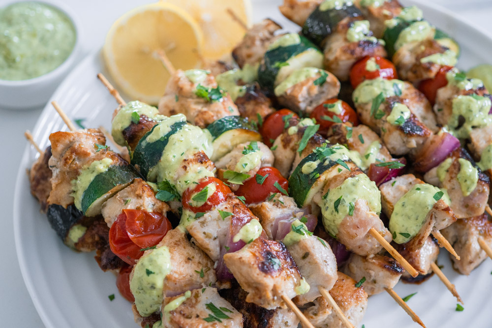 Mediterranean chicken skewers drizzled with pesto aioli on a white plate.